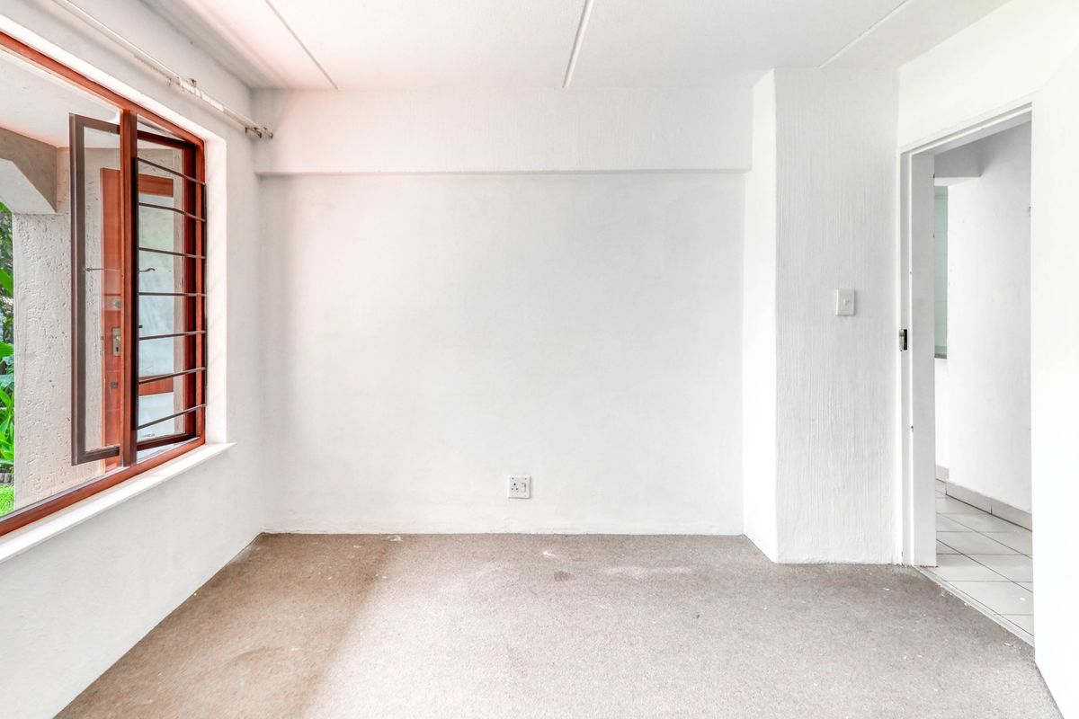 An empty bedroom featuring a large casement window with garden views, neutral walls, and carpeted flooring, ready for personalization.