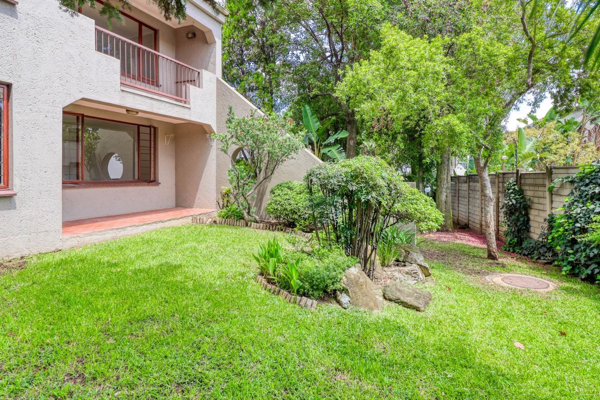A private rear garden featuring a landscaped lawn, established trees, and a covered patio area with balcony access above.