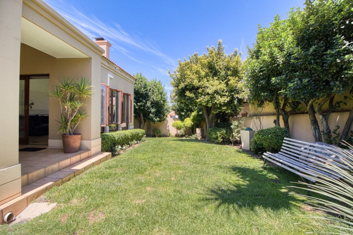 A well-maintained rear garden featuring a covered patio with tiled flooring, lush lawn, and established trees enclosed by a boundary wall.