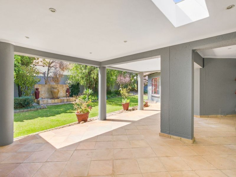 A spacious covered patio featuring tiled flooring, recessed lighting, a skylight, and structural columns, overlooking a landscaped garden with a built-in water feature.