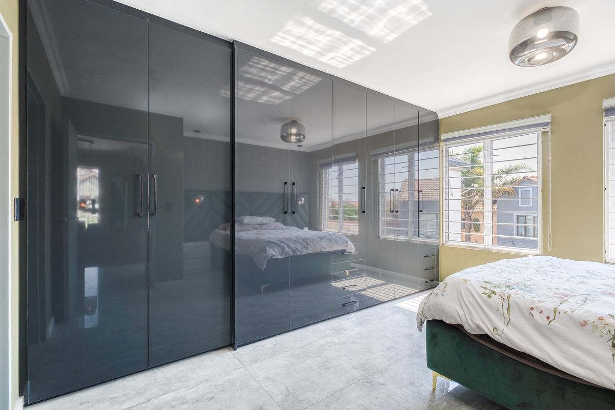 A modern bedroom featuring extensive floor-to-ceiling high-gloss built-in cupboards, tiled flooring, and large windows with security bars.
