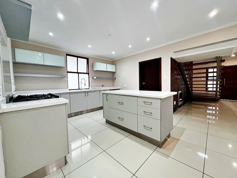 Modern kitchen featuring a central island, stone countertops, gas hob, and sleek cabinetry, seamlessly flowing into an open-plan area with a feature brick wall.