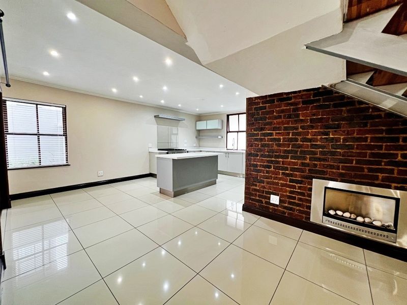 Spacious open-plan living and kitchen area featuring modern tiled floors, a central island, built-in fireplace, and an attractive exposed brick feature wall.