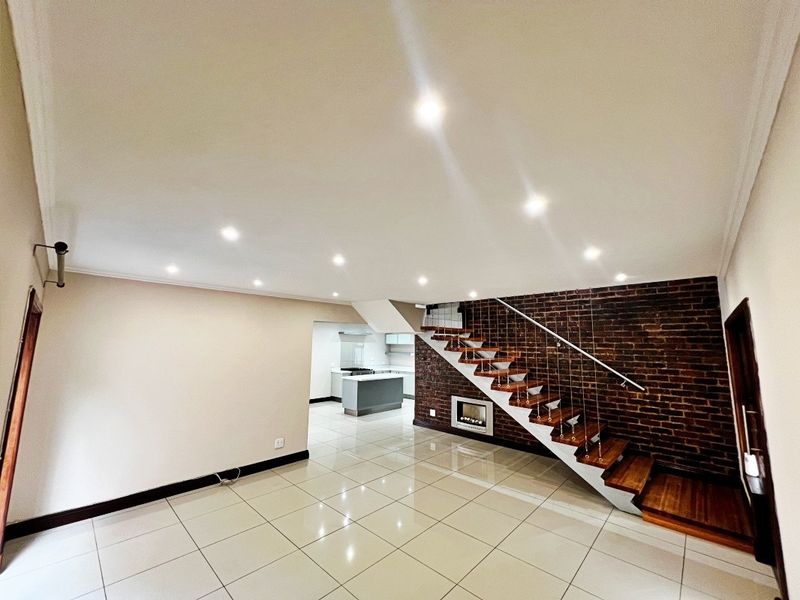 Spacious open-plan living area featuring polished porcelain tiles, an exposed brick wall, a modern floating staircase, and a sleek integrated kitchen.