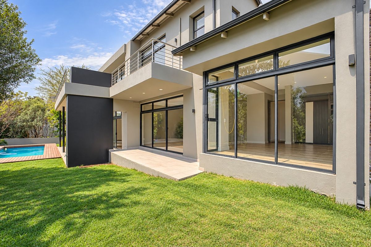 Rear view of a modern two-story house featuring a swimming pool, wooden deck, landscaped garden, and a spacious balcony with large sliding glass doors.
