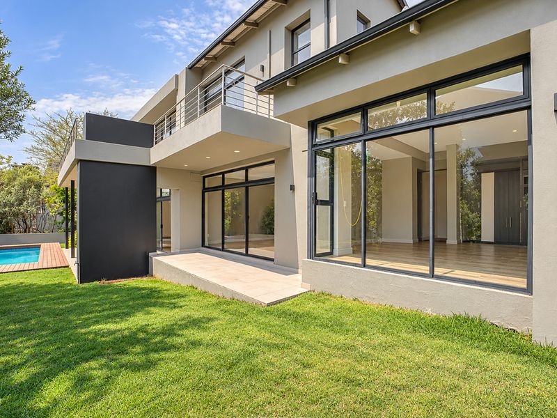 Rear view of a modern two-story house featuring a swimming pool, wooden deck, landscaped garden, and a spacious balcony with large sliding glass doors.