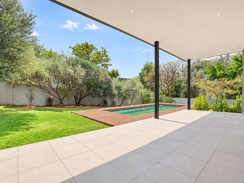 A modern covered patio featuring tiled flooring and recessed lighting, overlooking a swimming pool with timber decking and a well-maintained garden.