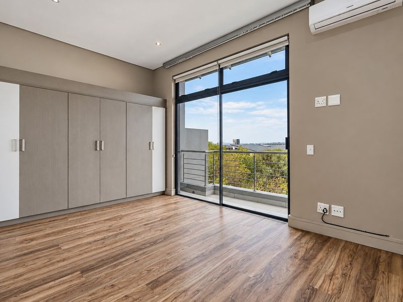 A bright, modern bedroom featuring built-in cupboards, laminate flooring, and a sliding glass door leading to a private balcony with scenic views.