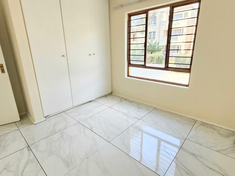 A bright, empty bedroom featuring modern marble-look tiled flooring, built-in cupboards, and a window fitted with security bars.