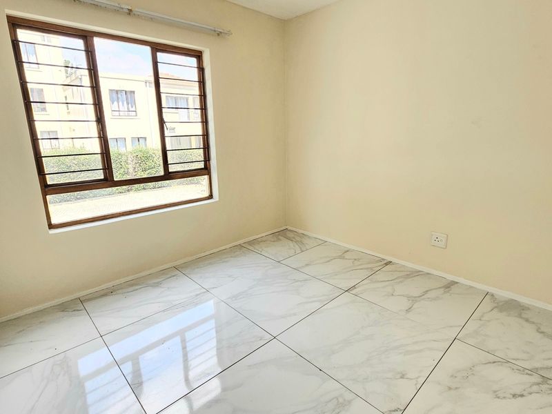 A bright, empty bedroom featuring modern marble-look floor tiles, a built-in cupboard, and a window fitted with security bars.