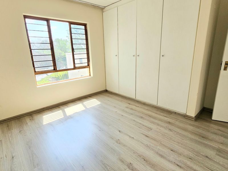 A bright, empty bedroom featuring light-toned laminate flooring, built-in storage cupboards, and a window fitted with security bars.