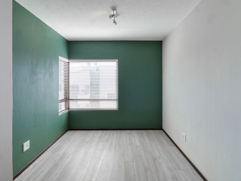 A bright, empty bedroom featuring modern light-toned laminate flooring, a corner window with fitted blinds, and a clean, contemporary green and white color scheme.