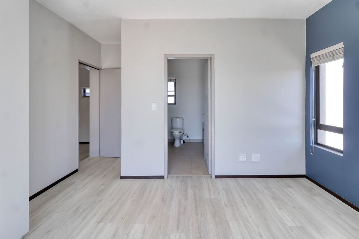 A bright, empty bedroom featuring light-toned laminate flooring, a blue accent wall, and direct access to an en-suite bathroom.