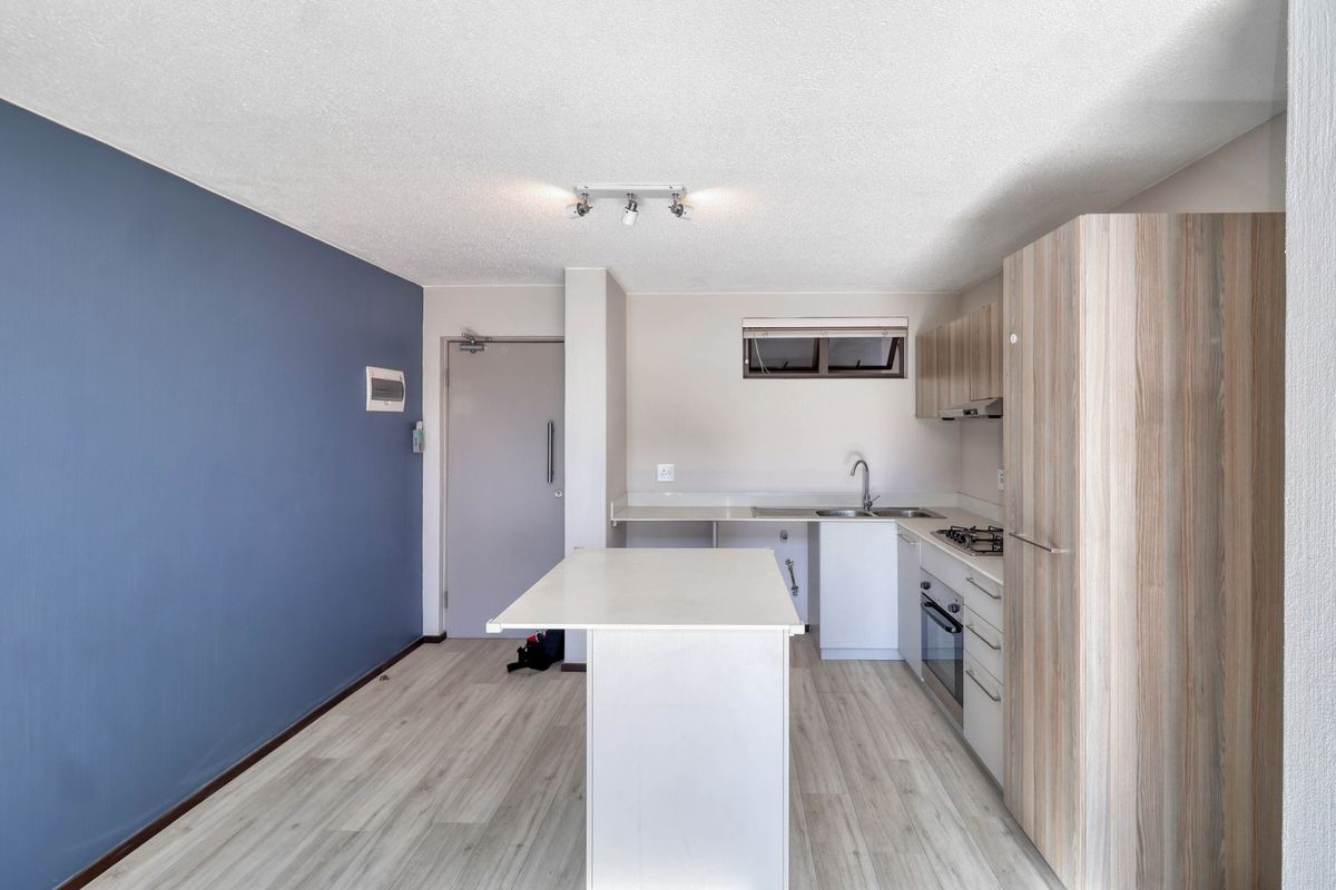 A modern, open-plan kitchen featuring light wood-tone cabinetry, a central island, built-in oven, gas hob, and a striking blue accent wall.