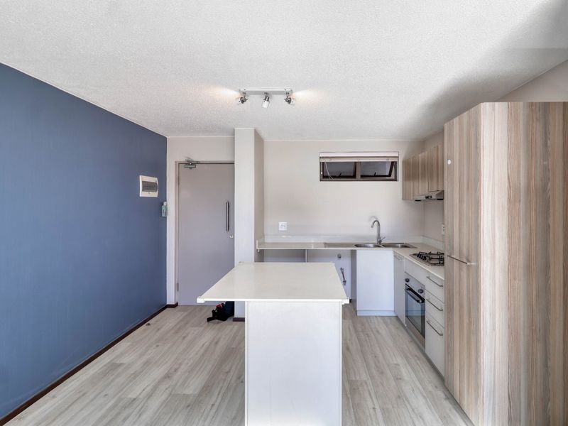 A modern, open-plan kitchen featuring light wood-tone cabinetry, a central island, built-in oven, gas hob, and a striking blue accent wall.