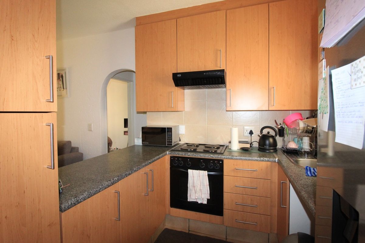 Compact kitchen featuring light wood-finish cabinetry, granite countertops, built-in oven, electric hob, and tiled backsplash with an arched doorway leading to the living area.