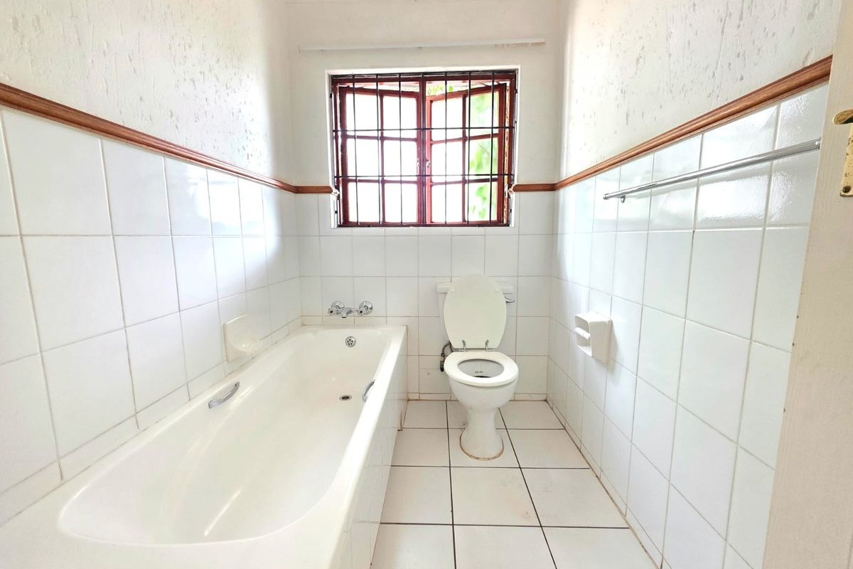 A dated bathroom featuring a standard bathtub, toilet, and white wall tiles with security bars on the window.