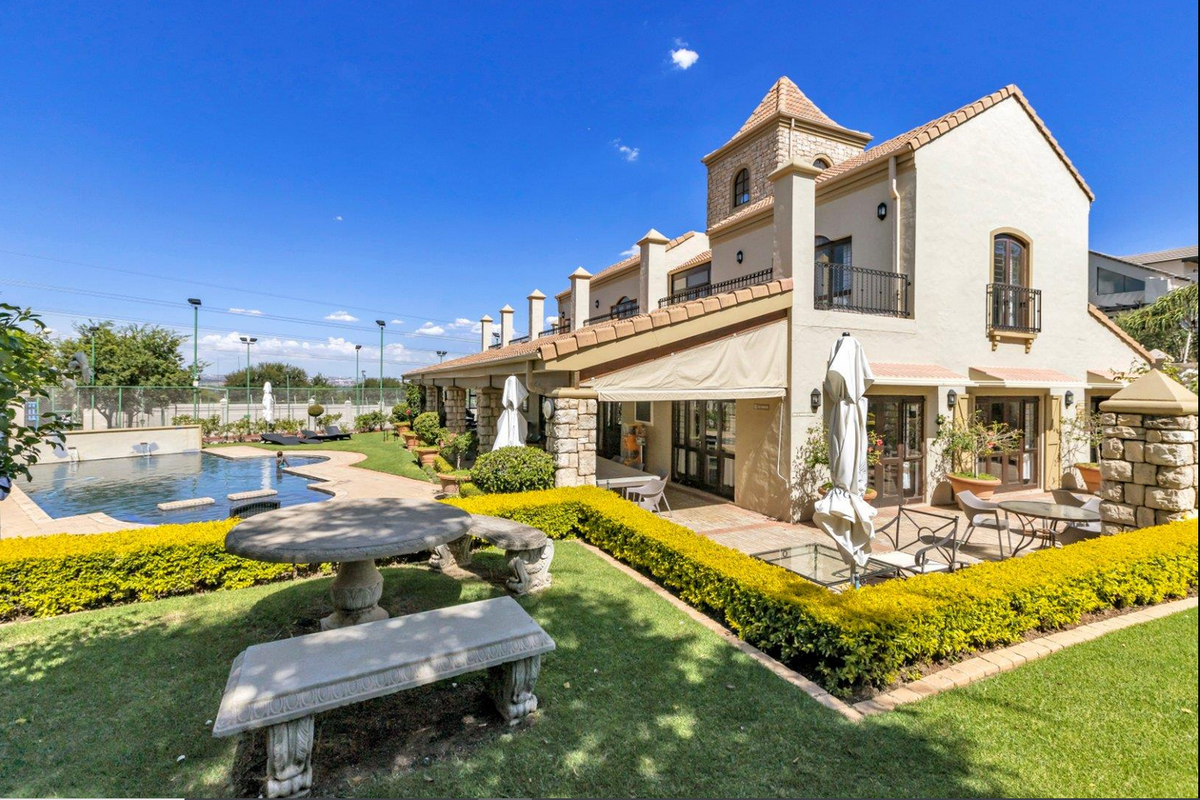 A well-maintained rear exterior view featuring a swimming pool, stone-paved patio, landscaped garden, and a multi-level building with a balcony and stone architectural details.