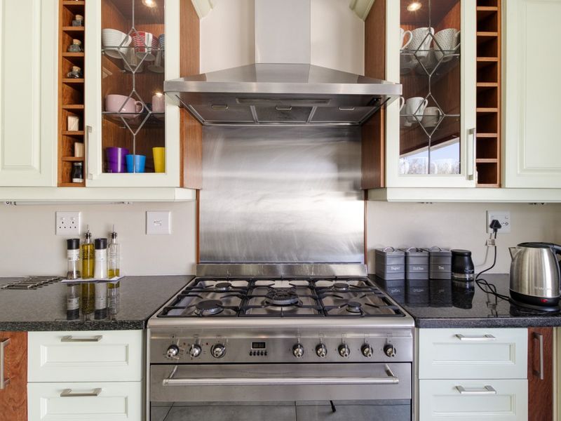 A well-maintained kitchen featuring a stainless steel gas stove, matching extractor fan, granite countertops, and elegant glass-fronted cabinetry with integrated wine storage.