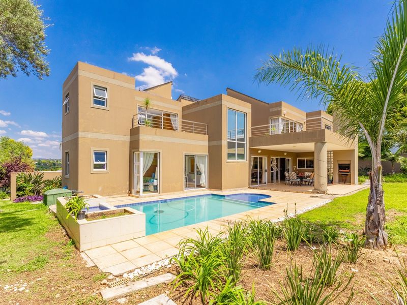 Rear view of a modern double-story house featuring a swimming pool, covered patio with built-in braai, and upper-level balconies overlooking the garden.