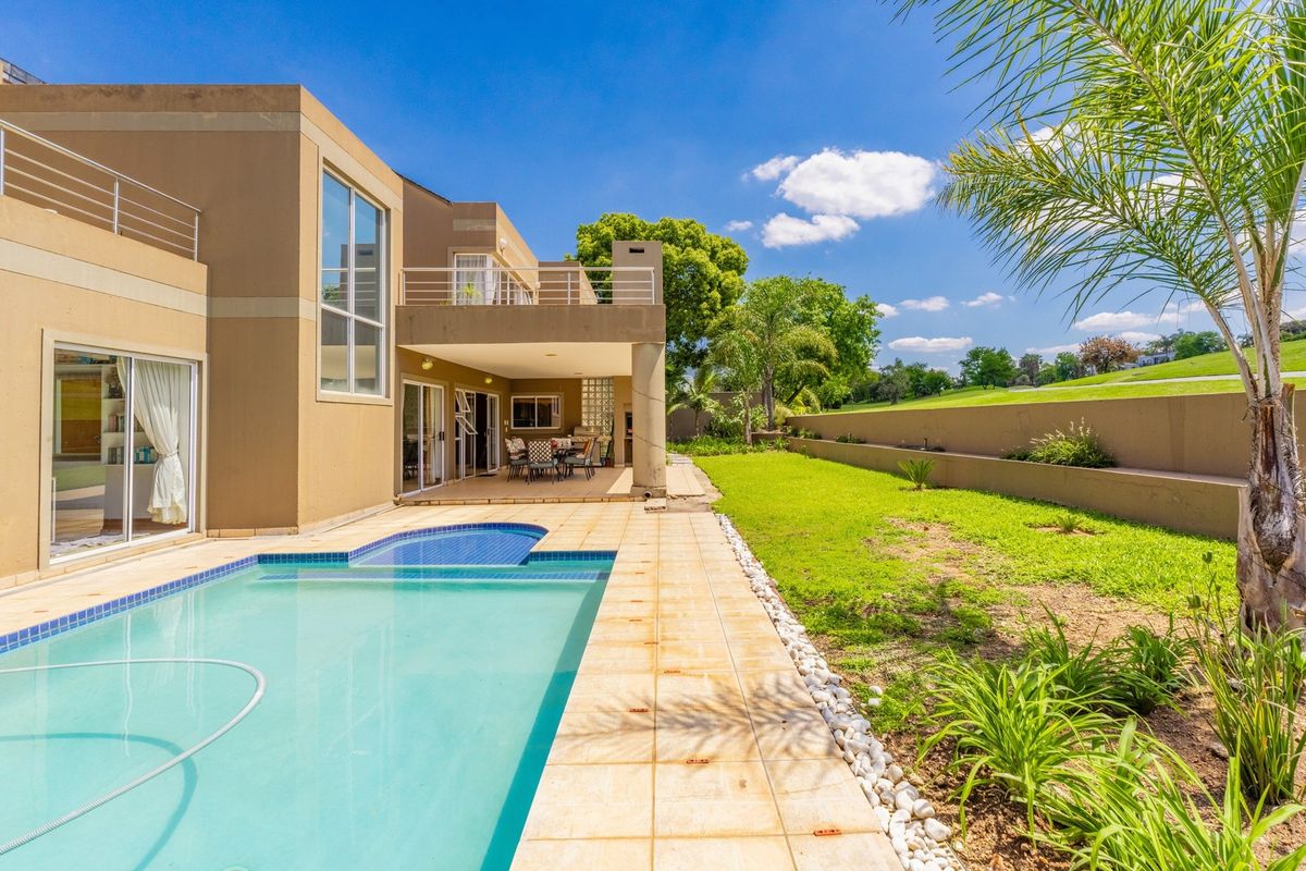 Rear view of the property featuring a swimming pool, covered patio, upper balcony, and a landscaped garden with a retaining wall.
