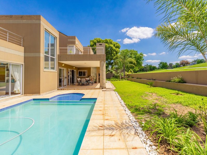Rear view of the property featuring a swimming pool, covered patio, upper balcony, and a landscaped garden with a retaining wall.