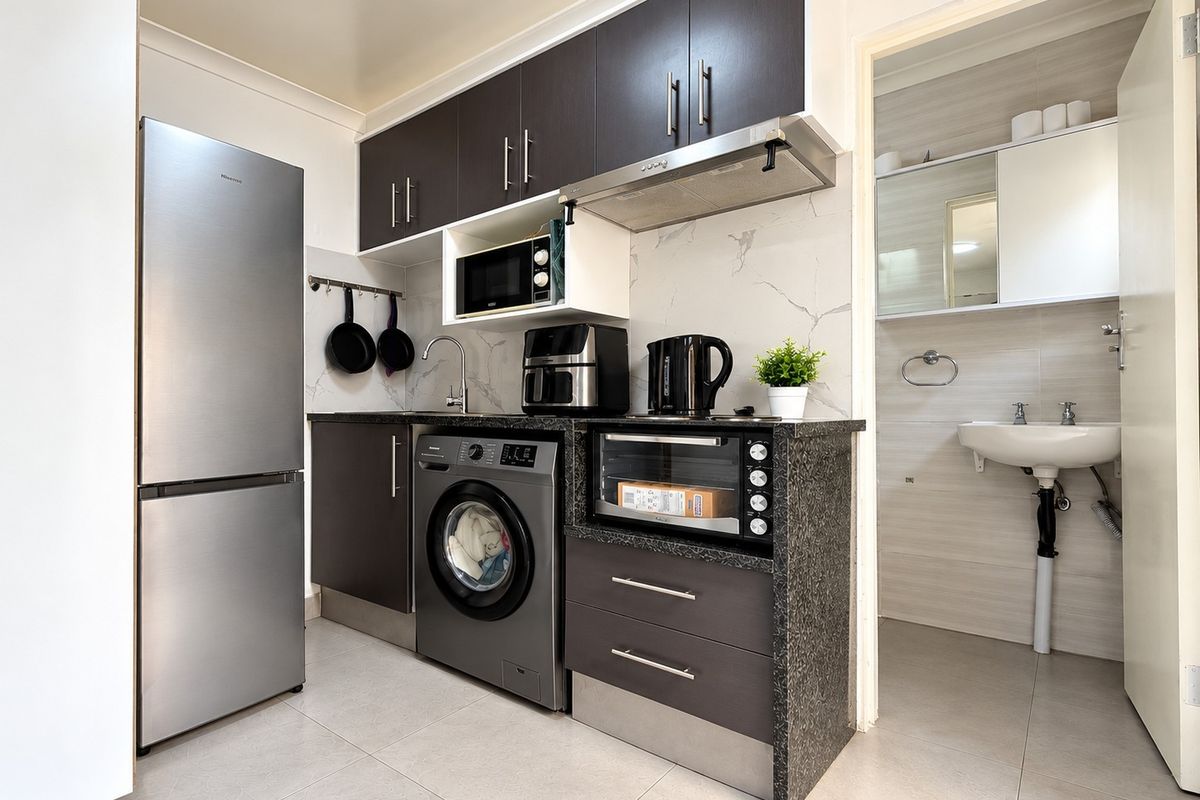 Compact kitchen featuring dark cabinetry, granite-look countertops, tiled backsplash, and integrated laundry plumbing, with an adjacent bathroom entrance.