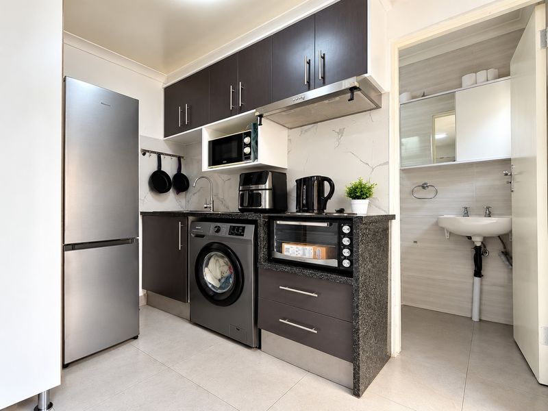 Compact kitchen featuring dark cabinetry, granite-look countertops, tiled backsplash, and integrated laundry plumbing, with an adjacent bathroom entrance.