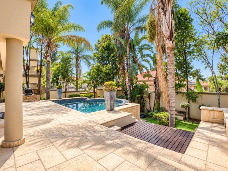 A well-maintained outdoor pool area featuring stone paving, timber decking, and a built-in braai, surrounded by established garden landscaping.