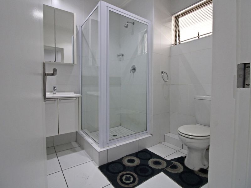 A clean, functional bathroom featuring a glass-enclosed shower, white vanity with basin, and a standard toilet, finished with neutral wall and floor tiles.
