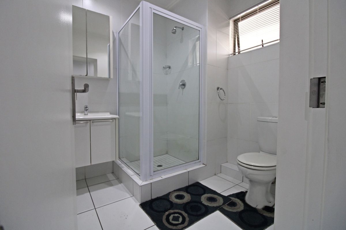 A clean, functional bathroom featuring a glass-enclosed shower, white vanity with basin, and a standard toilet, finished with neutral wall and floor tiles.