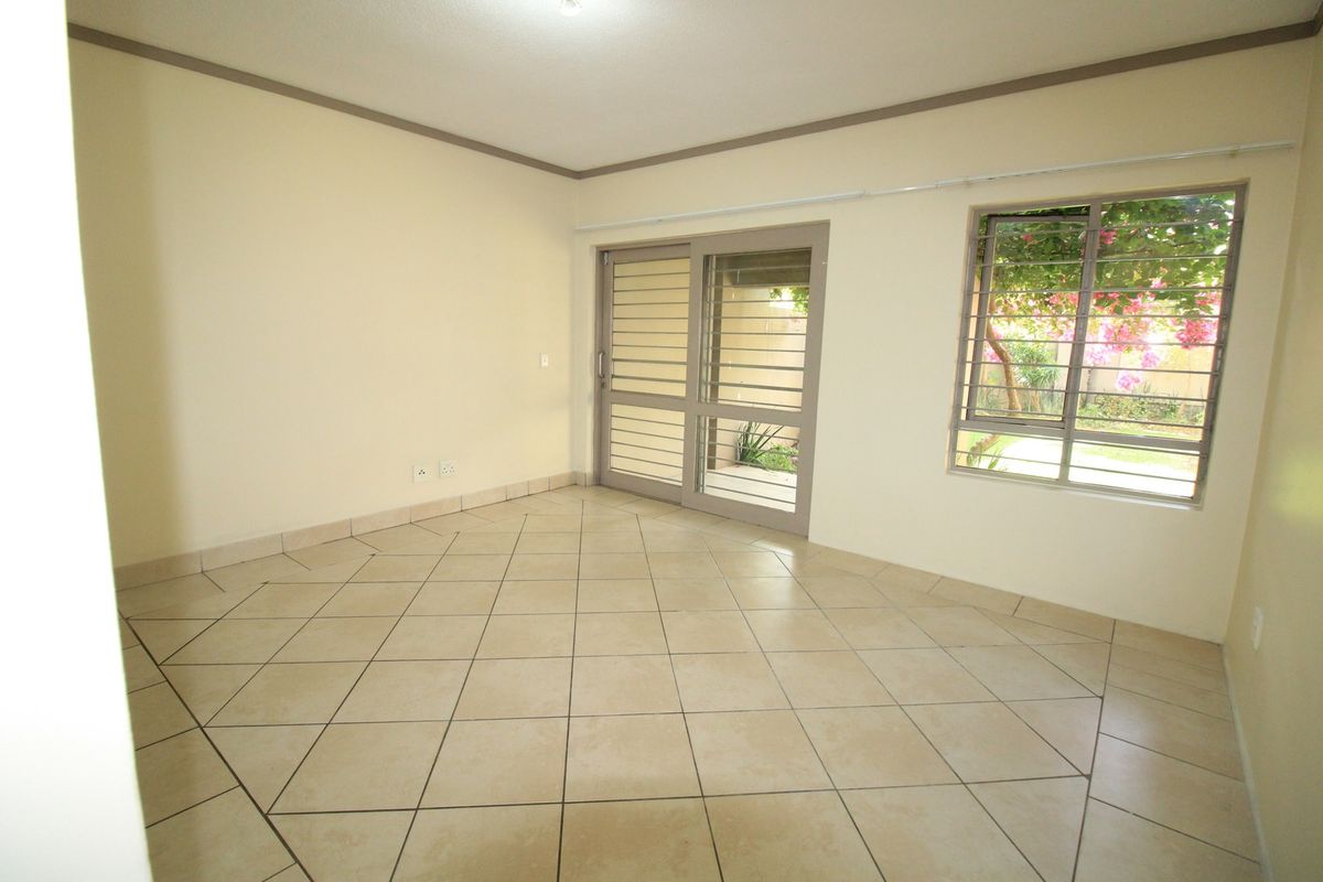 An empty living room featuring neutral tiled flooring, a sliding glass door with security bars, and a window overlooking the garden.