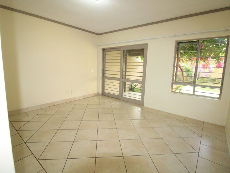 An empty living room featuring neutral tiled flooring, a sliding glass door with security bars, and a window overlooking the garden.