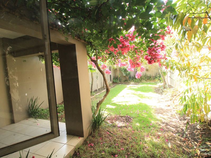 A private garden area featuring a small tiled patio, lush greenery, and a flowering bougainvillea tree, accessible via a sliding glass door.