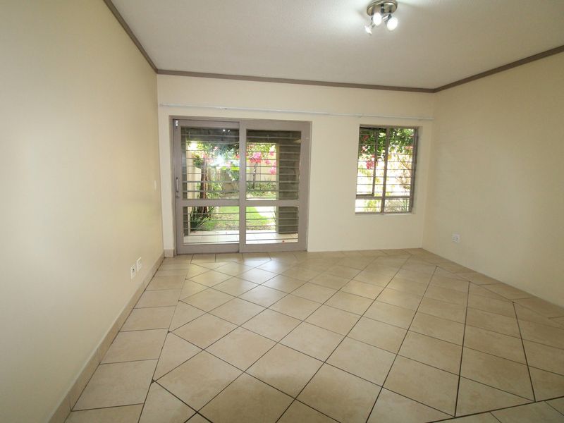 An empty living room featuring neutral tiled flooring, a sliding glass door with security gate, and natural light overlooking a garden area.
