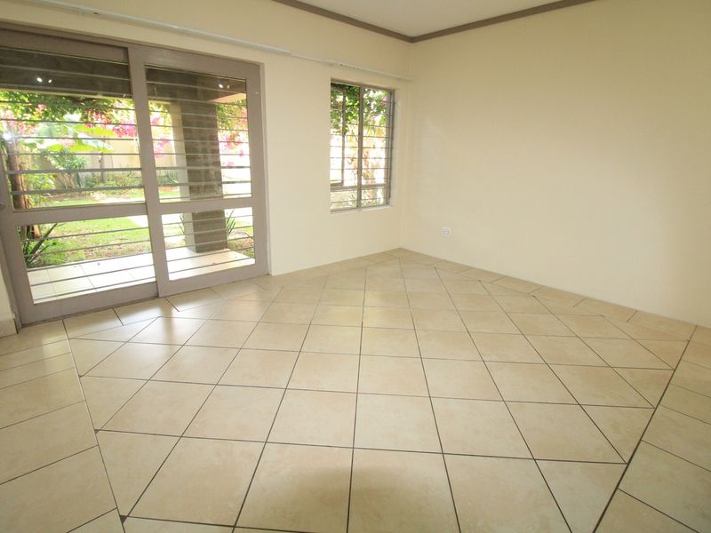 An empty, sunlit living room featuring neutral tiled flooring and a sliding glass door providing direct access to the garden area.