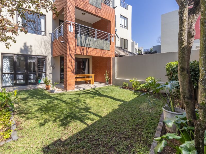 A private rear garden featuring a well-maintained lawn, boundary wall, and a covered patio area with brick detailing and a balcony above.