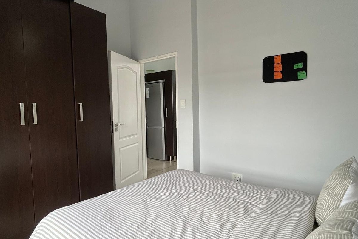A bedroom featuring a dark wood built-in wardrobe and neutral wall finishes, with a view into an adjacent kitchen area.
