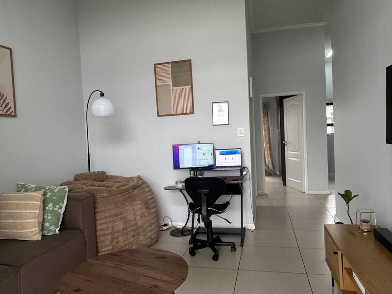 A living area featuring neutral tiled flooring and access to an adjacent hallway, providing a functional space for relaxation or home office use.