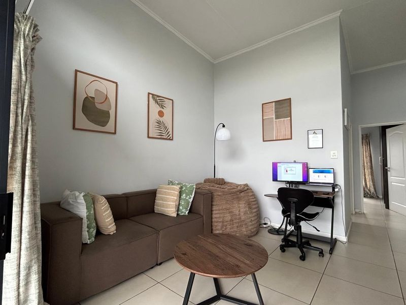 A bright living area featuring neutral tiled flooring and high ceilings, currently utilized as a combined lounge and home office space.