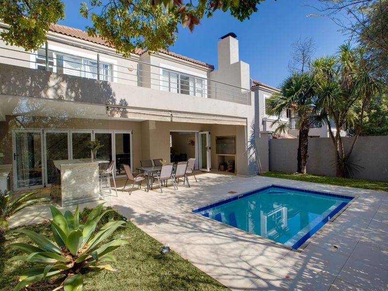 A well-maintained rear patio area featuring a swimming pool, built-in braai, and a spacious balcony above with stainless steel balustrades.