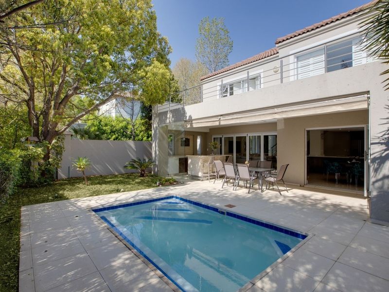 Rear exterior view featuring a swimming pool, tiled patio area with a built-in bar, and a balcony overlooking the garden.