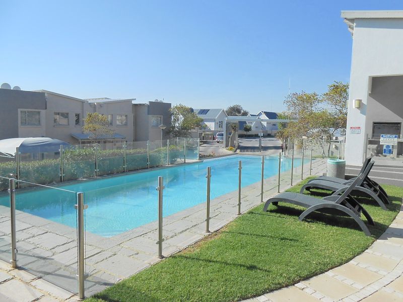 A well-maintained communal swimming pool area featuring glass safety fencing, paved surrounds, and a manicured lawn section within an apartment complex.