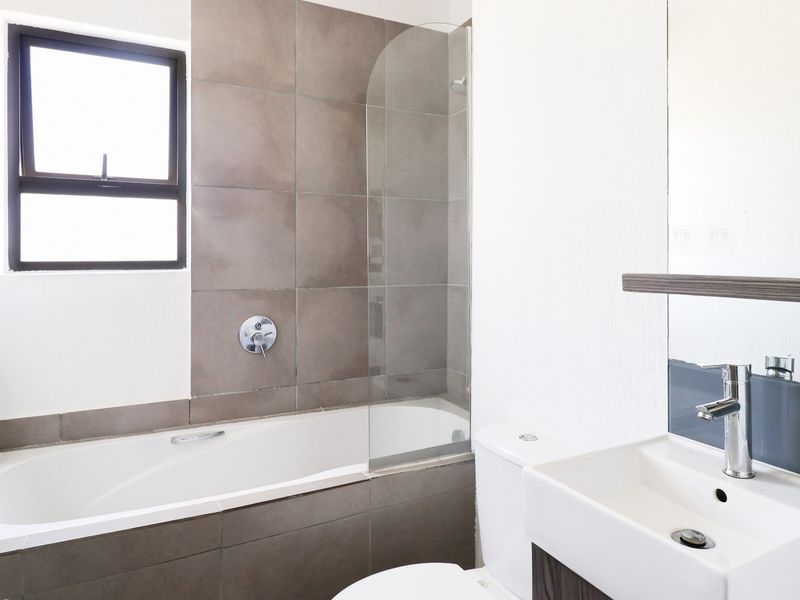 A clean, modern bathroom featuring a built-in bathtub with a glass shower screen, a vanity basin, and a toilet with neutral wall tiling.