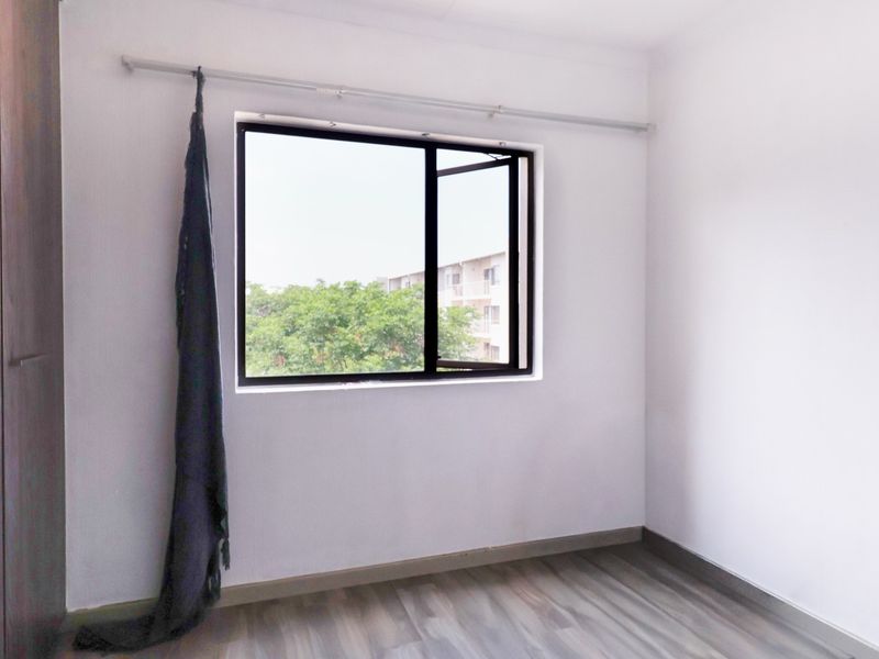 A bright, empty bedroom featuring modern laminate flooring, a built-in wardrobe, and an aluminium-framed window with a curtain rod.