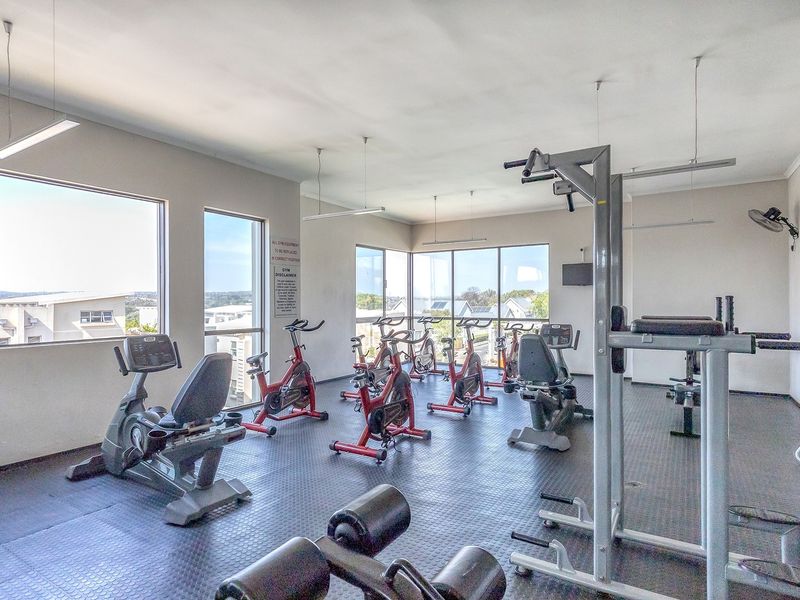 A well-lit apartment complex gym featuring rubberized flooring, large windows with natural light, and various fixed exercise equipment.