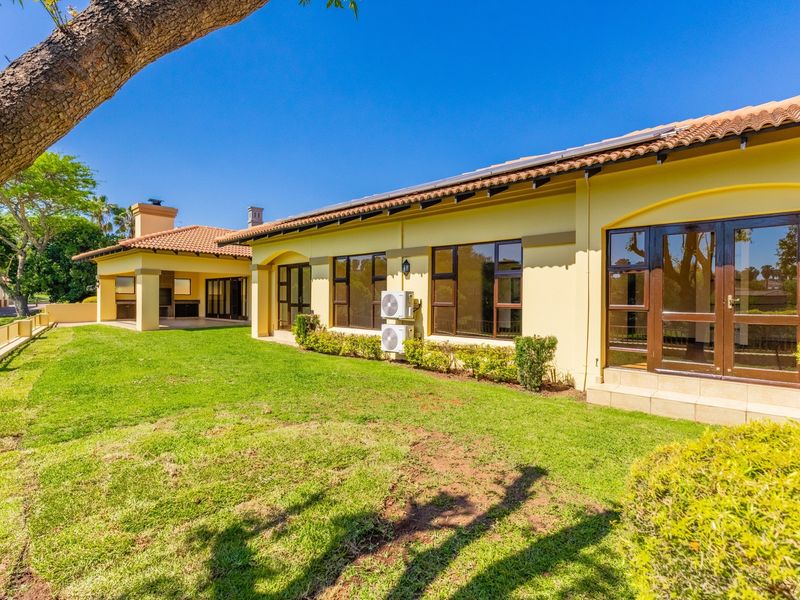 Spacious rear exterior view featuring a well-maintained lawn, covered patio with built-in braai, and classic terracotta roof tiles.