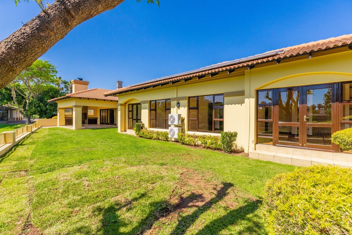 Spacious rear exterior view featuring a well-maintained lawn, covered patio with built-in braai, and classic terracotta roof tiles.