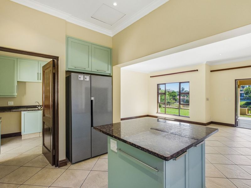 Spacious open-plan kitchen area featuring a central island with granite countertops, adjacent scullery, and tiled flooring, flowing into a bright living space.