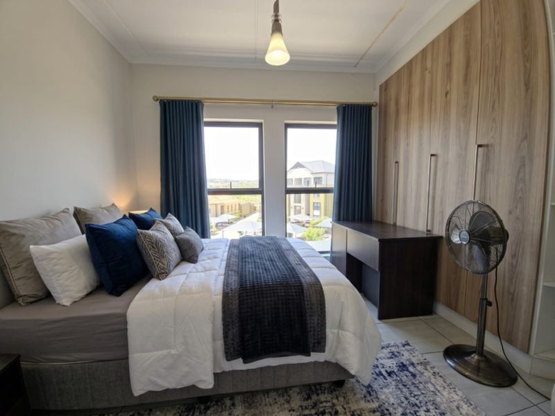 A well-maintained bedroom featuring built-in wooden cupboards, tiled flooring, and large windows that provide natural light and views of the surrounding complex.